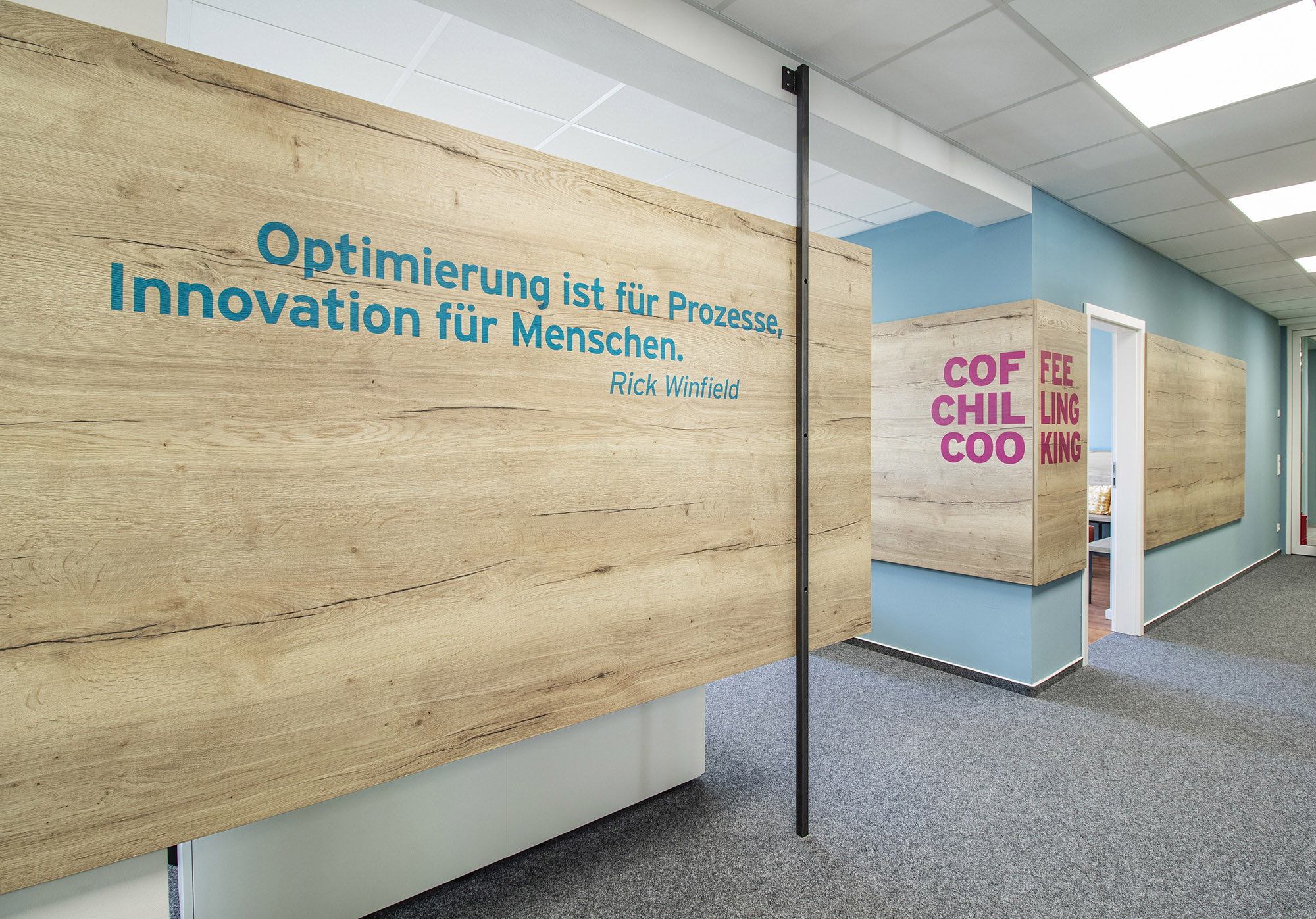 Ein partieller Raumtrenner im gleichen Format wie die Wandverkleidung im Hintergrund trägt das Zitat "Optimierung ist für Prozesse, Innovation für Menschen."