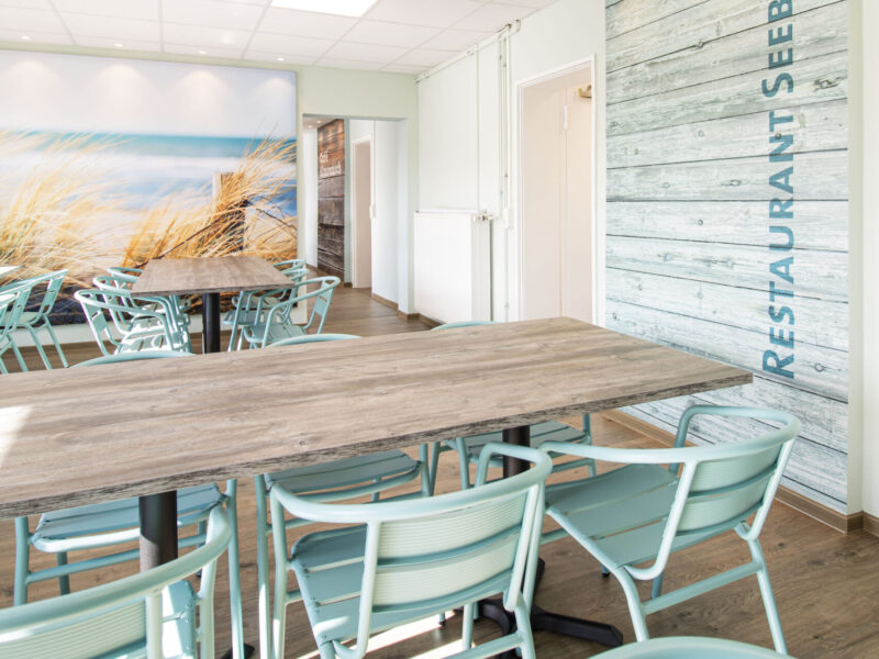 Ansicht der Kantine im Strandthema. Im Hintergrund der Durchgang zu Kantine Almrausch und Dünenmotiv. Im Vordergrund Esstische in Holzoptik, mit Grün-cyan farbenen Stühlen. Auf der rechten Raumseite eine Holzverkleidung mit Beschriftung, Restaurant.