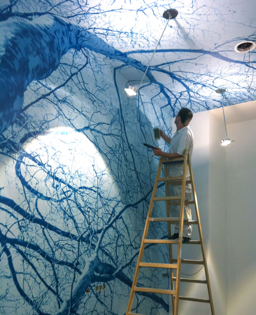 Ein Maler steht auf einer hohen Leiter und klebt eine Bildtapete mit einem blau eingefärbtem Baummotiv an Wand und Decke.