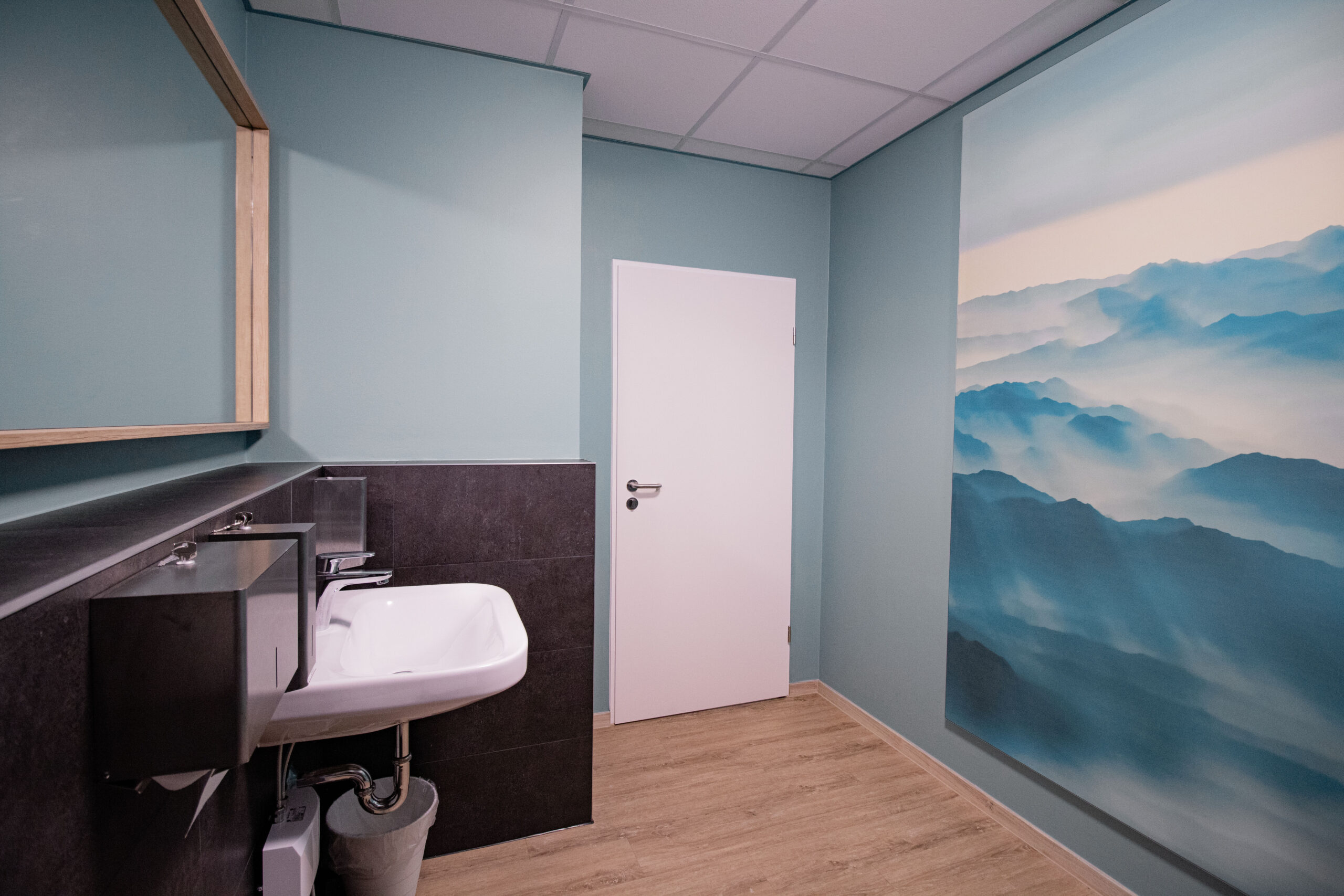 Patienten WC mit Fußboden in Holzoptik. Cyan-Blau gestrichene Wände. Auf der linken Bildseite ein weißes Waschbecken, hängt an einer Vorsatzschale mit grauen Fliesen. Darüber ein Spiegel mit Holzrahmen. Gegenüberliegend hängt ein Akustikbild mit nebligem Bergmotiv an der Wand.