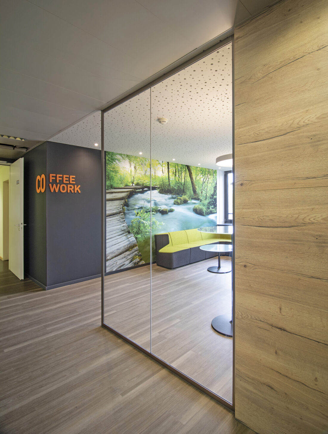 An der Raumecke von Flur zu Lounge ist das Wortspiel COffee und COwork in einem kräftigen Orange auf grauer Wand zu sehen. Durch die Glaswand sieht man die Motivtapete, die einen Wald mit Bachlauf und Steg zeigt. Davor ist ein Sofamöbel, mit farbiger Sitzfläche und Lehne platziert.