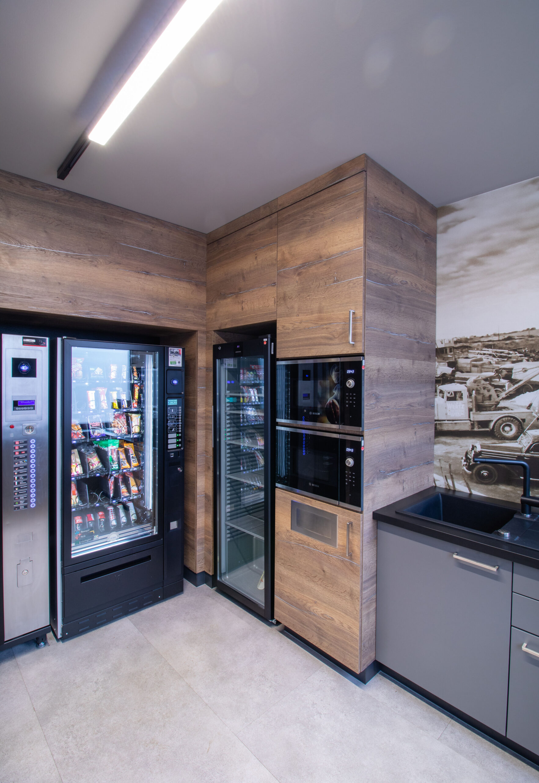 Hochschränke und Einlassungen mit integrierten Getränke-Automaten, Snack-Automaten und Mikrowellengeräten. Rechts ein Stück der Motivtapete mit historischem Schwarz-Weiß-Bild.