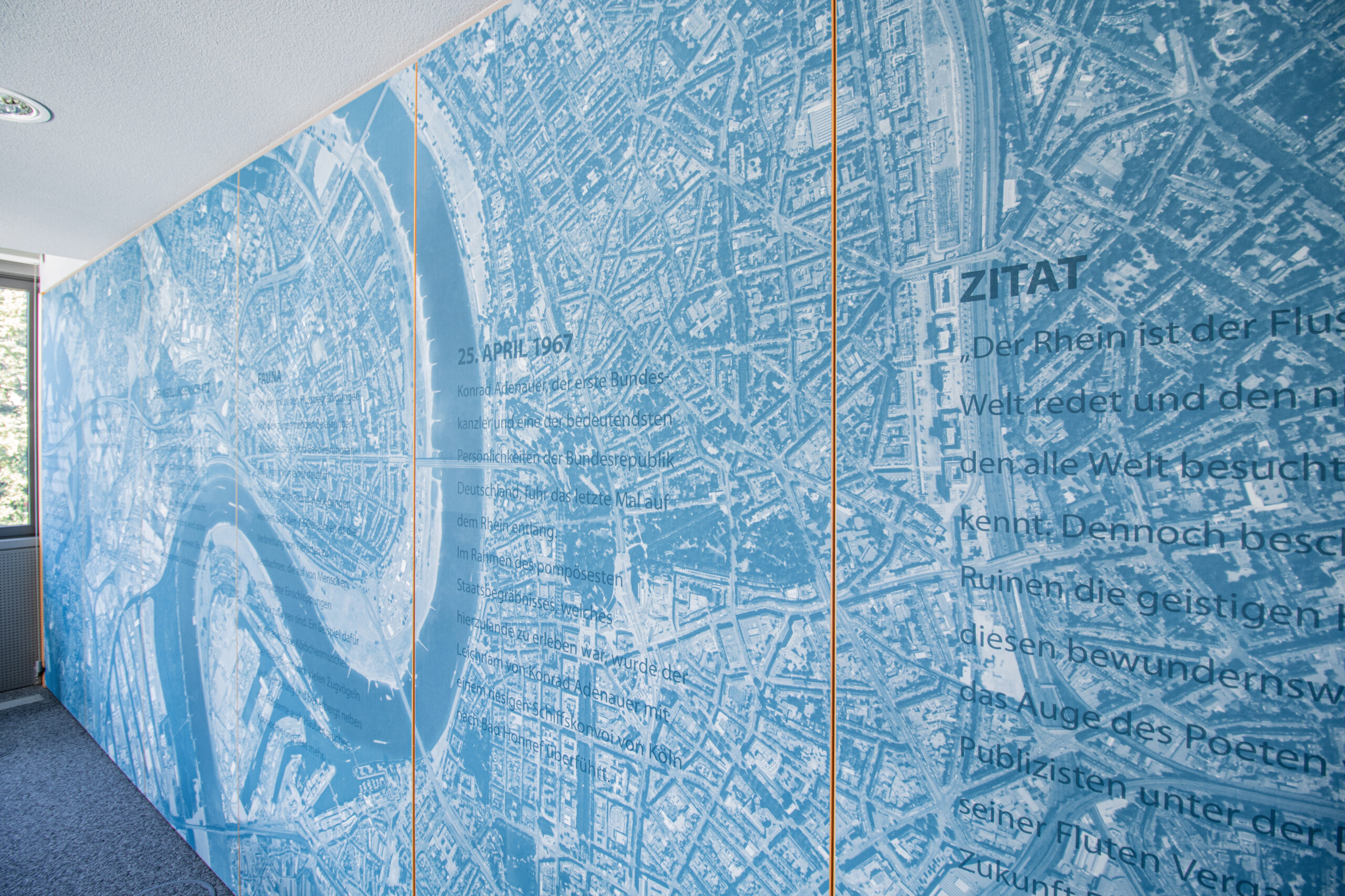 Eine Luftaufnahme der Stadt Köln mit Flusslauf des Rheins, ist in einem Blauton eingefärbt und als Motivtapete verarbeitet. Auf den einzelnen Tapetenbahnen sind verschiedene Schriftblöcke, z.B mit Informationen über den Niebelungenschatz, die Fauna und ein Zitat über den Rhein, aufgeführt.