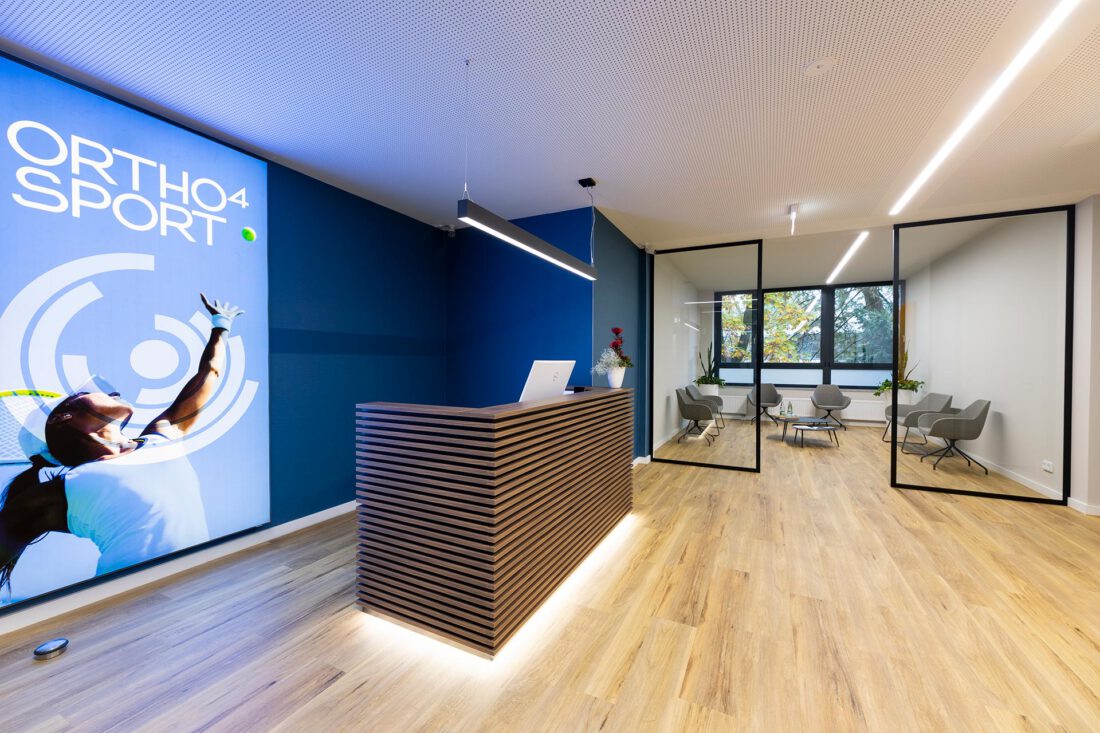 Empfangsbereich einer modern gestalteten Praxis mit Holzlamellen-Tresen, blauem Wanddesign, Glaswänden und lichtdurchflutetem Wartebereich, perfekt geplant für eine ansprechende Praxisgestaltung.
